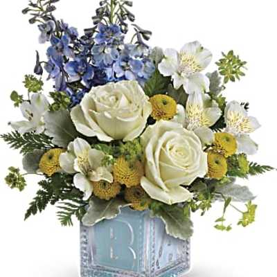 Low arrangement of white roses, blue delphinium, and yellow button flowers in a blue baby block glass cube
