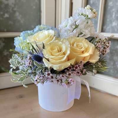 Yellow roses and blue hydrangea in a white hat box with pink filler flowers.