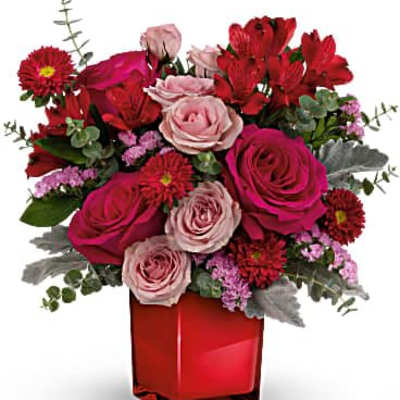 Short red and pink flower arrangement with roses and other blooms in a red cube vase