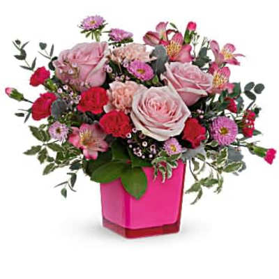 Pink and red roses, carnations, and mixed flowers arranged in a bright pink square vase