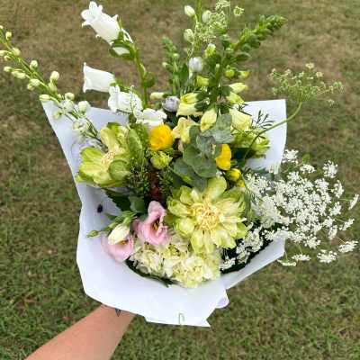 Handheld bouquet of yellow, white, and pink mixed flowers wrapped in white paper over a grassy lawn