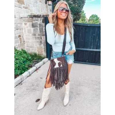 Woman wearing sunglasses, a light top, denim shorts, and white boots with a fringe crossbody bag.