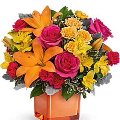 Bright arrangement of orange lilies, pink roses, and yellow blooms in an orange cube vase