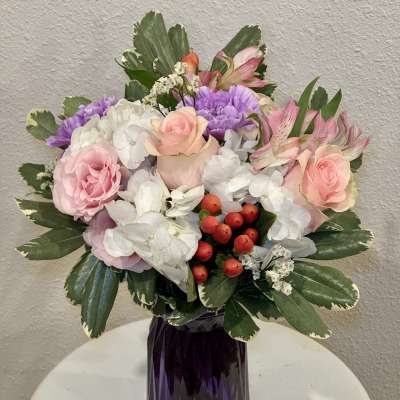 Pink and white mixed bouquet with purple accents and red berries in a faceted dark purple glass vase