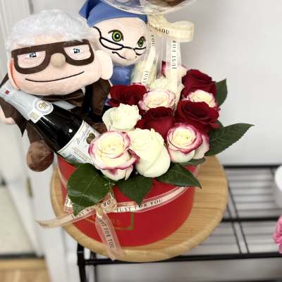 Gift set with red and white roses in a red box, two plush dolls, a bottle, and a clear anniversary balloon.