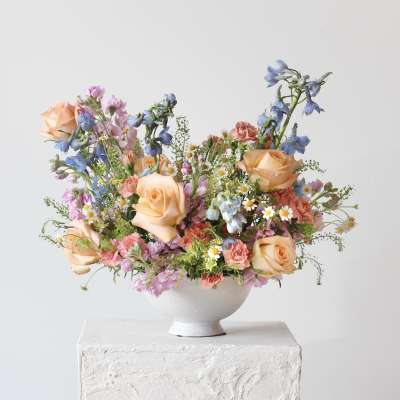 Mixed bouquet of peach roses, pink blooms, and blue flowers in a white bowl vase