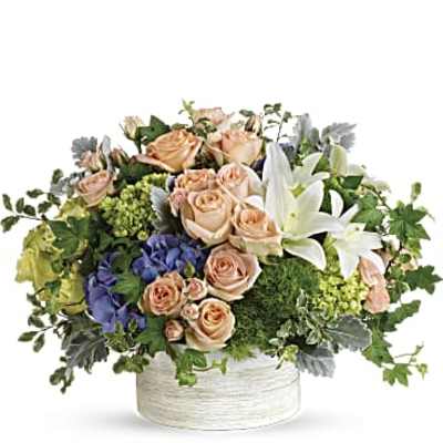 Low arrangement of peach roses, white lilies, and blue hydrangeas in a white ceramic container