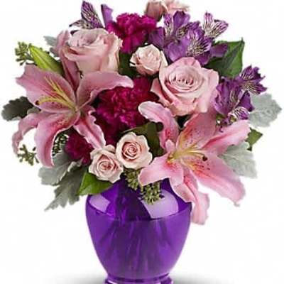 Pink lilies, roses, and purple blooms arranged in a bright purple glass vase