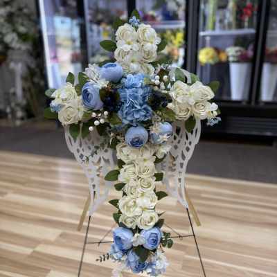 Tall cross-shaped arrangement of white and light blue roses with angel wing accents on a metal stand.