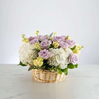 Low basket arrangement with lavender roses, white hydrangeas, and soft yellow flowers.