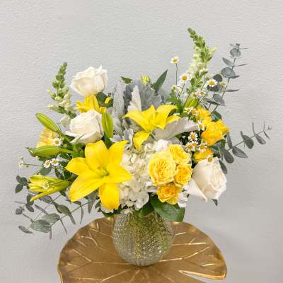 Yellow lilies and roses with white roses and hydrangea in a clear textured vase with silver foliage.
