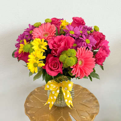 Bright mixed bouquet of pink, purple, and yellow flowers in a glass vase with a yellow ribbon.