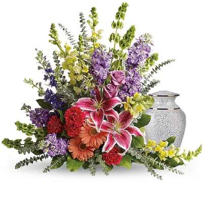Colorful floral arrangement with lilies, roses, and gerbera daisies beside a white urn