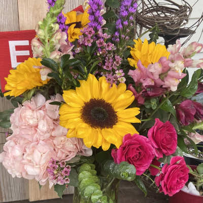 Mixed bouquet with sunflowers, pink roses, hydrangea, and purple spikes in a glass vase