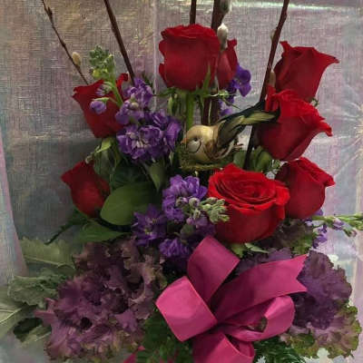 Red roses and purple flowers in a tall glass vase with a pink ribbon
