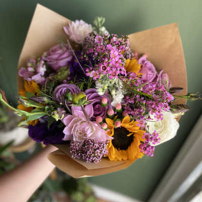 Handheld bouquet of pink, purple, white, and yellow flowers wrapped in brown paper