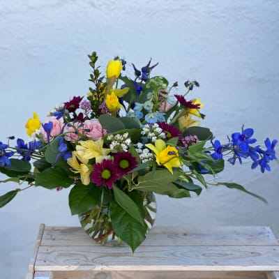 Mixed bouquet in a clear glass vase with blue, yellow, pink, and purple flowers.