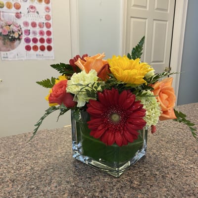 Mixed bouquet in a square glass vase with red, orange, yellow, and pale green blooms