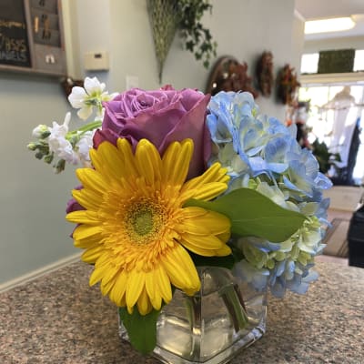 Colorful bouquet with a yellow gerbera daisy, purple rose, and blue hydrangea in a glass vase