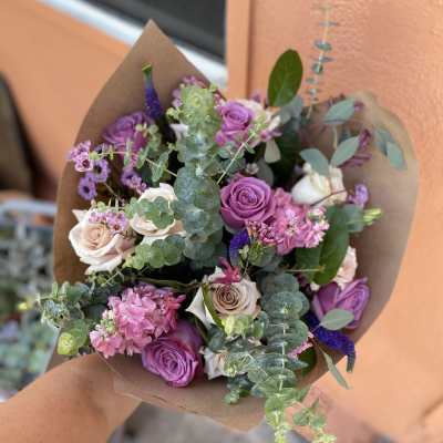 Handheld bouquet of purple and blush roses with eucalyptus in brown paper