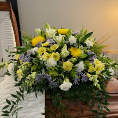 Large floral spray on a wooden casket with yellow, white, and lavender flowers.