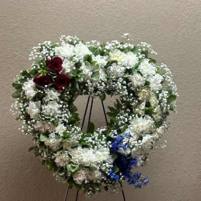 Heart-shaped floral wreath with white flowers, red roses, and blue accents on a stand