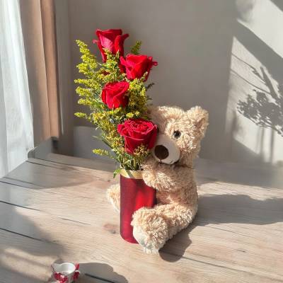 Red roses in a red vase beside a teddy bear