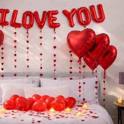 Red heart balloons and "I LOVE YOU" balloons above a bed with candles and heart confetti.