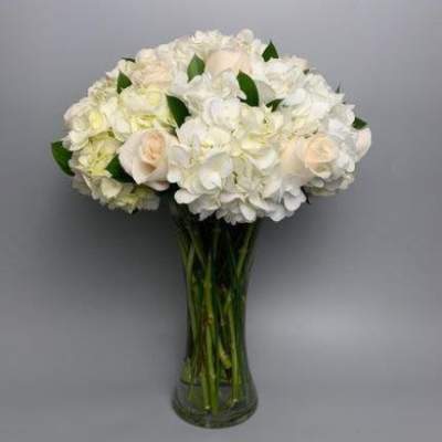Tall clear glass vase filled with white hydrangeas and ivory roses in a rounded arrangement
