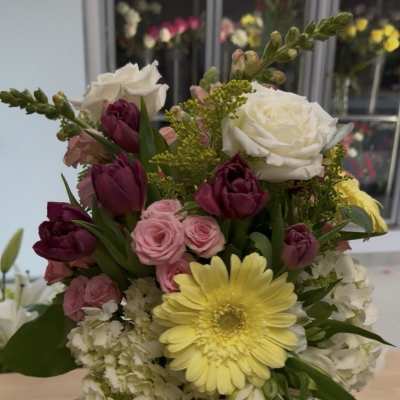 Mixed bouquet of yellow gerbera, white roses, pink spray roses, and dark tulips in a clear glass vase
