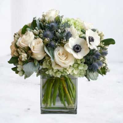 Low glass vase of white roses, white anemones, green hydrangeas and blue accent blooms