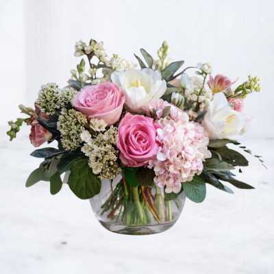 Low vase arrangement of pink roses, white blooms, and blush hydrangea in a clear glass bowl.