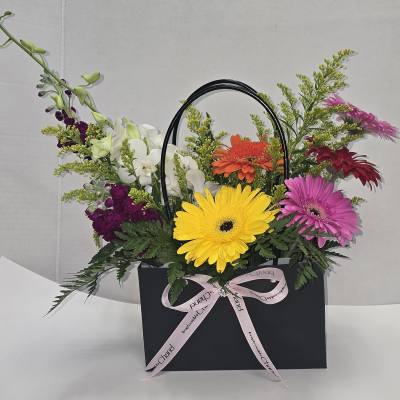 Mixed gerbera and orchid arrangement in a black gift bag with a pink ribbon