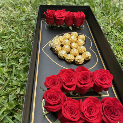 Black gift box with red roses and gold-wrapped chocolates on grass