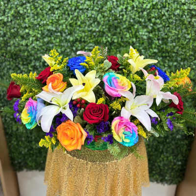 Low arrangement of rainbow and red roses with white and yellow lilies on a gold cloth.