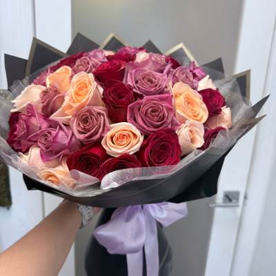 Bouquet of pink, peach, and red roses wrapped in black paper with a lavender ribbon