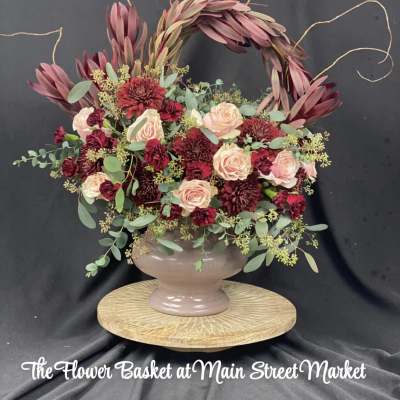 A burgundy and blush floral arrangement in a pedestal vase with a decorative arch.