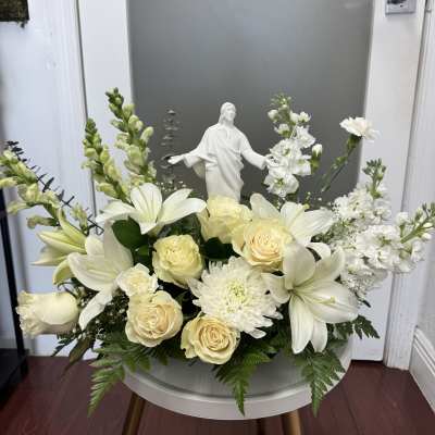 White floral arrangement with roses, lilies, and a small religious statue