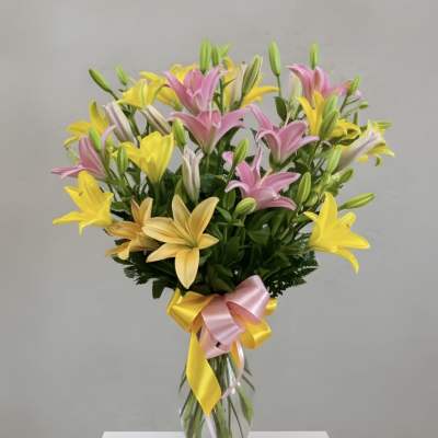 Tall clear vase of pink and yellow lilies with buds, tied with a pink and yellow ribbon bow.