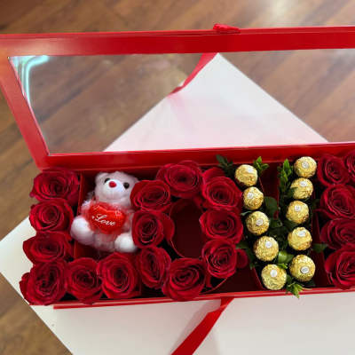 Red roses arranged in a red gift box with a small teddy bear and chocolates