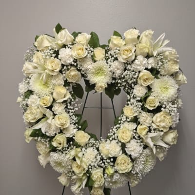Heart-shaped standing wreath of white roses, lilies, and mums on a metal easel.