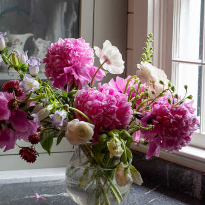 Loose arrangement of pink peonies and white blooms in a clear glass vase on a countertop.