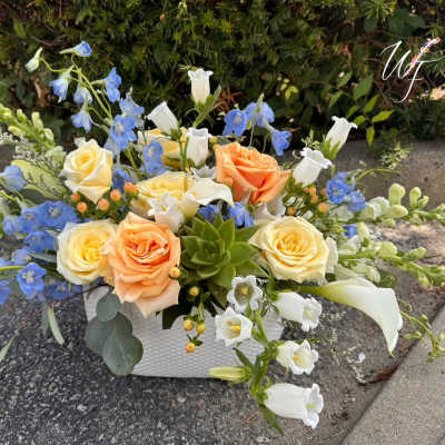 Mixed bouquet of roses, blue delphinium, and white bell-shaped flowers in a white container