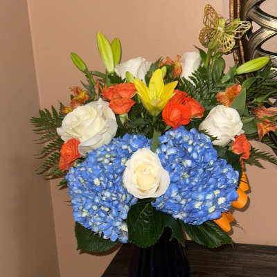 Bouquet of blue hydrangeas, white roses, orange roses, and yellow lilies in a dark vase
