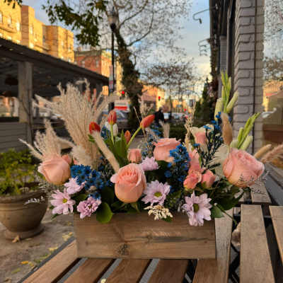 Low rustic box arrangement with peach roses, blue asters, and pastel mums on an outdoor café table.