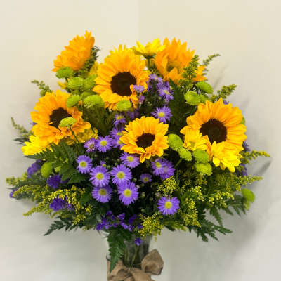 Tall arrangement of yellow sunflowers and purple asters in a clear vase with a burlap bow