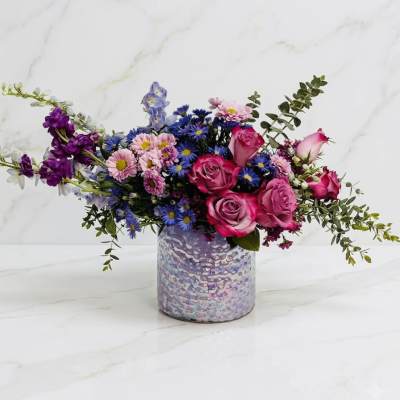 Asymmetrical arrangement of pink roses and purple and blue blossoms in an iridescent lavender vase