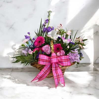 Basket arrangement of pink gerbera daisies, lavender roses, and purple flowers with a pink bow.
