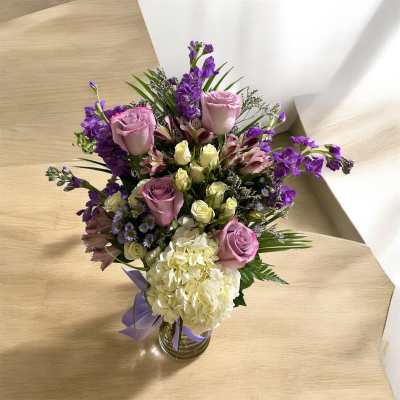 Tall mixed bouquet of pink roses, purple blooms, and white hydrangea in a clear glass vase