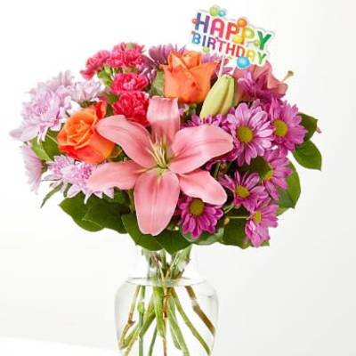 Mixed pink and orange flowers in a clear glass vase with a birthday topper